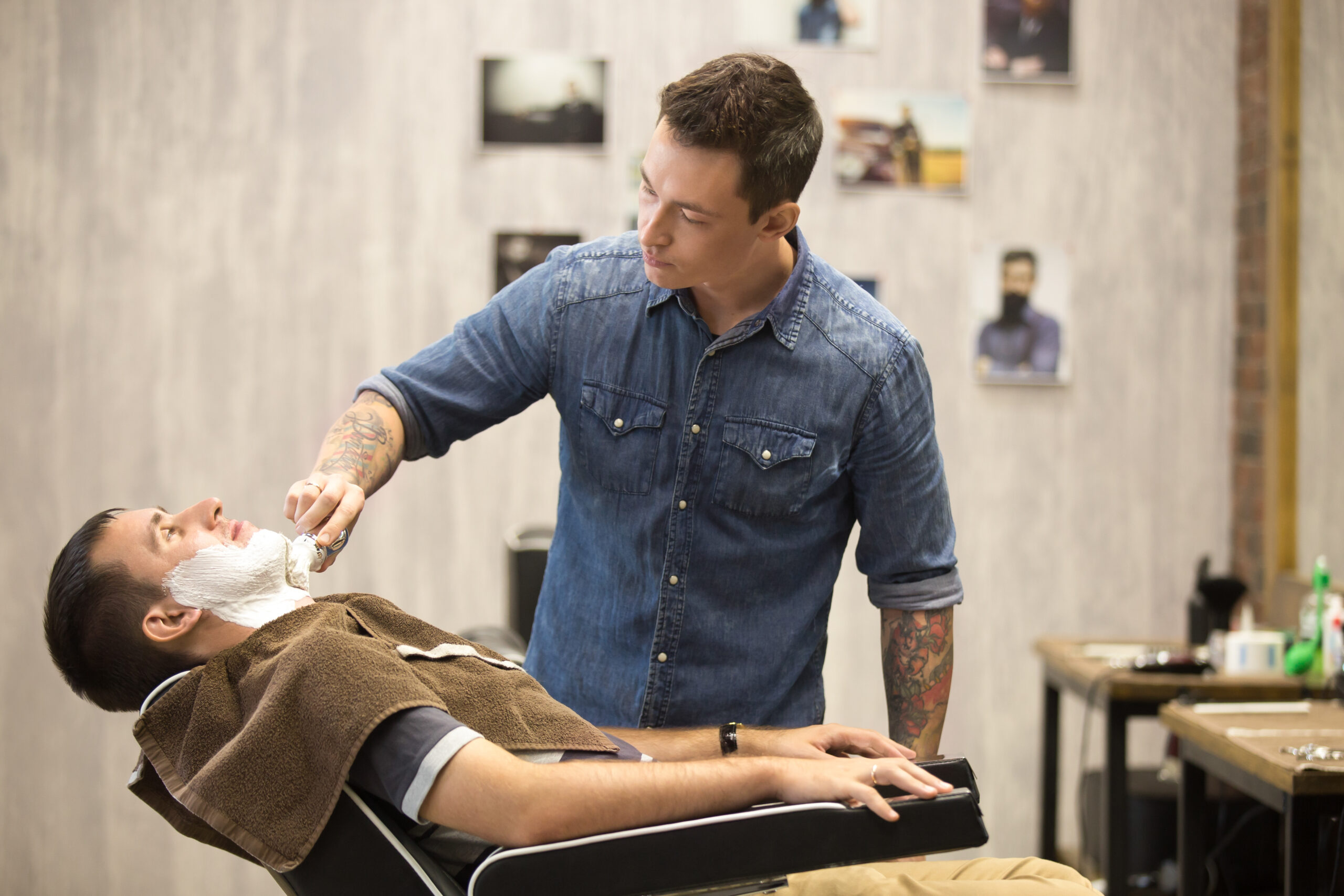 Interior shot of working process in modern barbershop. Side view portrait of handsome young man getting beard shaving. Male hairdresser serving client, putting shaving soap before using straight razor